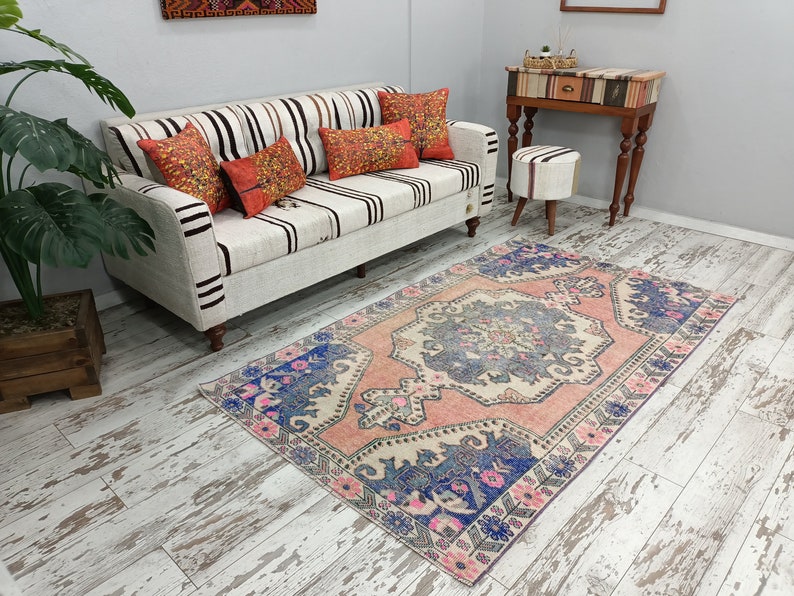 Pode incluir: Um tapete vintage rosa e azul com padr&atilde;o sobre fundo branco. O tapete tem um design floral e est&aacute; colocado sobre um piso de madeira branqueada em frente a um sof&aacute; branco com almofadas listradas.