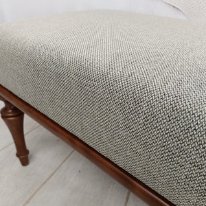 May include: A light grey fabric ottoman with a dark wood frame and turned legs. The ottoman has a textured fabric surface.