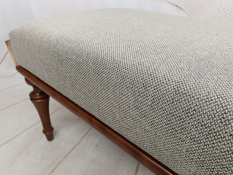 May include: A light grey fabric ottoman with a dark wood frame and turned legs. The ottoman has a textured fabric surface.