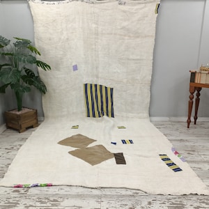May include: A white rug with a patchwork design featuring geometric shapes in brown, yellow, blue, and purple. The rug is laid out on a wooden floor.