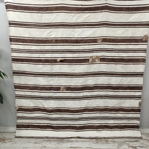 Puede incluir: Una alfombra crema con rayas horizontales marrones de diferentes anchos. La alfombra tiene varios parches cuadrados peque&ntilde;os de un material texturizado con dibujos. La alfombra est&aacute; colgada contra una pared gris claro.