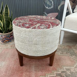 Peut inclure: Un pouf rond avec un dessus à motifs rouges et crème, des côtés tissés crème et des pieds en bois brun foncé. Le pouf est posé sur un tapis rose. Une plante en pot et une chaise blanche sont en arrière-plan.