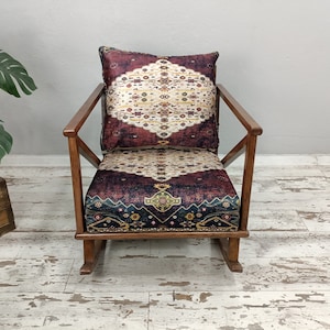 May include: A wooden rocking chair with a patterned cushion and backrest. The cushion and backrest feature a burgundy, cream, and navy blue design. The chair has a dark wood frame and is set on a light-colored wooden floor.