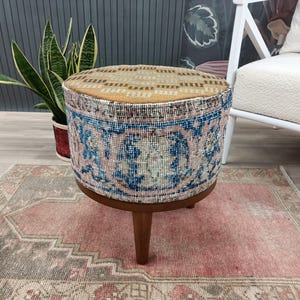 Può includere: Pouf rotondo con piano e lati in tessuto fantasia. I lati presentano un motivo floreale blu, rosa e crema. Il pouf ha una base in legno e tre gambe in legno. Sotto il pouf c'è un tappeto rosa e grigio.