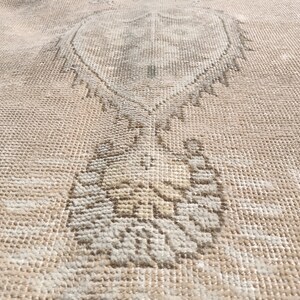 May include: A close-up of a beige rug with a faded, intricate design. The design features a central motif that resembles a stylized tree or plant.