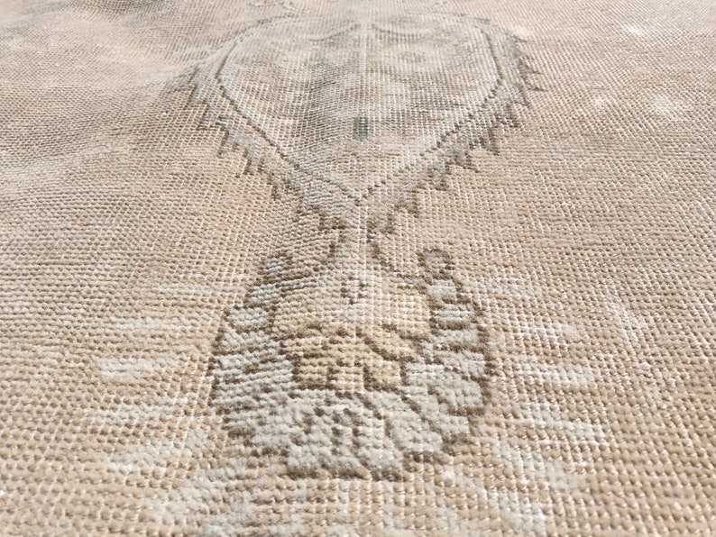 May include: A close-up of a beige rug with a faded, intricate design. The design features a central motif that resembles a stylized tree or plant.