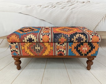 Op de afbeelding: Een rechthoekse opbergottoman met een kleurrijke geometrische stoffen bovenkant en houten poten. De stof heeft een mix van oranje, rood, blauw en zwarte kleuren.