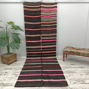 Puede incluir: Dos alfombras largas y rayadas con fondo marrón y rayas rosas, rojas y blancas. Las alfombras están colgadas en una pared y se encuentran en una habitación con paredes blancas y suelo de madera.