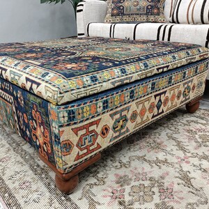 May include: A square ottoman with a blue, orange, and cream patterned fabric top and wooden legs. The ottoman is sitting on a cream-colored rug with a floral pattern.