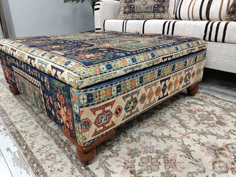 May include: A square ottoman with a blue, orange, and cream patterned fabric top and wooden legs. The ottoman is sitting on a cream-colored rug with a floral pattern.