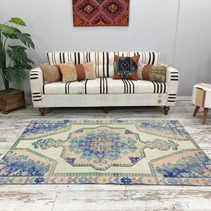 K&ouml;nnte beinhalten: Ein hellblauer und rosafarbener Vintage-Teppich mit Blumenmuster. Der Teppich liegt auf einem wei&szlig; get&uuml;nchten Holzboden vor einem wei&szlig;en Sofa mit gestreiftem Bezug und Kissen.