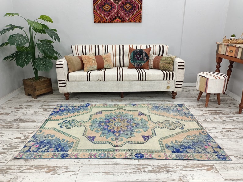 K&ouml;nnte beinhalten: Ein hellblauer und rosafarbener Vintage-Teppich mit Blumenmuster. Der Teppich liegt auf einem wei&szlig; get&uuml;nchten Holzboden vor einem wei&szlig;en Sofa mit gestreiftem Bezug und Kissen.