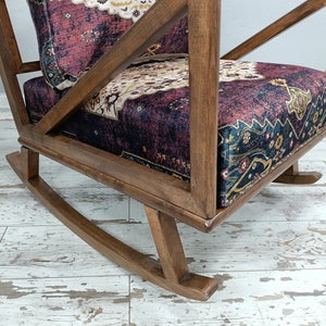 May include: A wooden rocking chair with a patterned cushion. The chair has a dark wood frame and a seat cushion with a burgundy, cream, and navy pattern. The chair's design includes a slatted back and curved rockers.