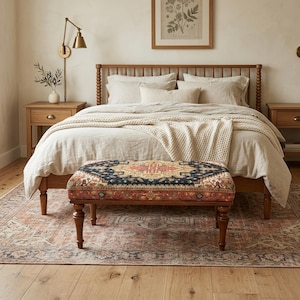a cozy and inviting bedroom with a large bed, wooden headboard, and a colorful patterned ottoman. The room has a warm and rustic atmosphere, with a wooden floor and a framed picture on the wall.