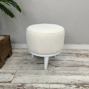 Puede incluir: Otomana redonda blanca con una cubierta de tela blanca esponjosa y patas de madera blancas.