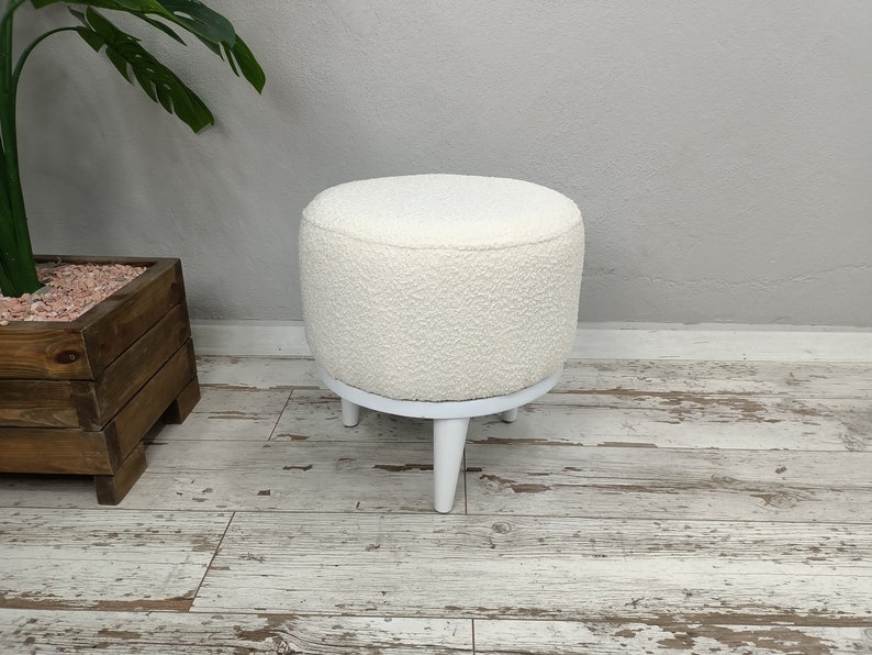 Puede incluir: Otomana redonda blanca con una cubierta de tela blanca esponjosa y patas de madera blancas.