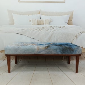 Puede incluir: Un banco de madera con un asiento de tela de estampado abstracto azul y marr&oacute;n. El banco est&aacute; en un dormitorio con una cama blanca y una manta blanca.