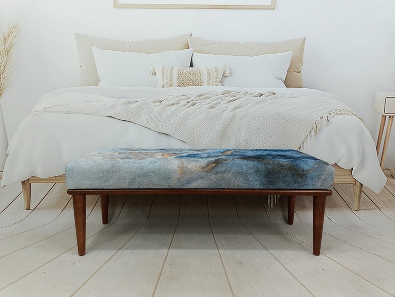 Puede incluir: Un banco de madera con un asiento de tela de estampado abstracto azul y marr&oacute;n. El banco est&aacute; en un dormitorio con una cama blanca y una manta blanca.