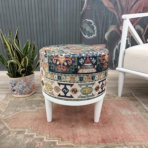 a colorful, patterned ottoman placed on a rug, with a potted plant and a white chair visible in the background.