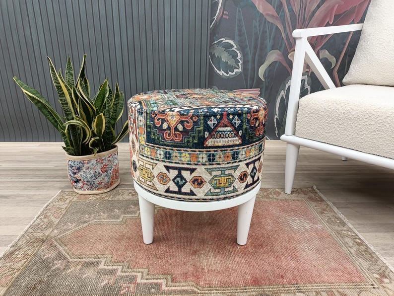 a colorful, patterned ottoman placed on a rug, with a potted plant and a white chair visible in the background.