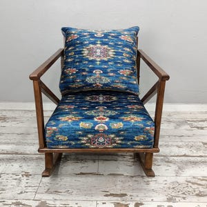 A blue and gold patterned chair with a matching pillow sits against a white wall on a wooden floor.