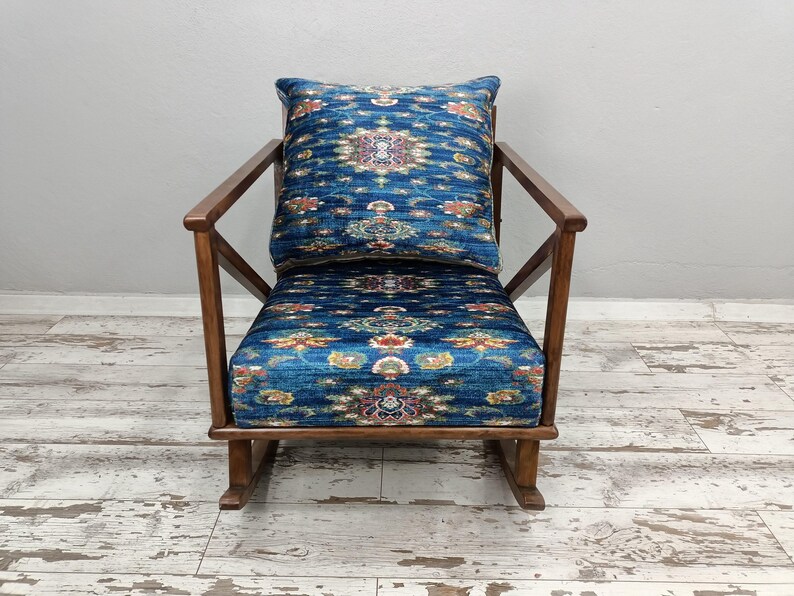 A blue and gold patterned chair with a matching pillow sits against a white wall on a wooden floor.