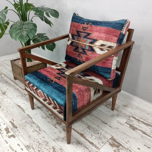 K&ouml;nnte beinhalten: Holzsessel mit geometrisch gemustertem Kissen und R&uuml;ckenlehne. Das Kissen und die R&uuml;ckenlehne weisen ein Design mit roten, blauen und wei&szlig;en Farben auf. Der Stuhl hat ein dunkles Holzgestell und steht vor einer Pflanze.