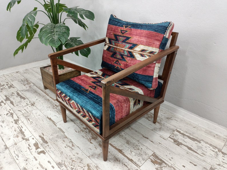 K&ouml;nnte beinhalten: Holzsessel mit geometrisch gemustertem Kissen und R&uuml;ckenlehne. Das Kissen und die R&uuml;ckenlehne weisen ein Design mit roten, blauen und wei&szlig;en Farben auf. Der Stuhl hat ein dunkles Holzgestell und steht vor einer Pflanze.