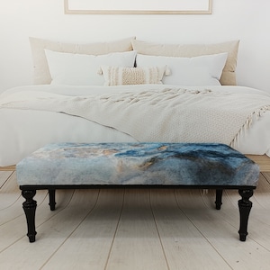 Puede incluir: Un banco con estampado azul y gris con patas de madera negra. El banco est&aacute; en un dormitorio con una cama blanca y una manta blanca.