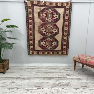 May include: A vintage Turkish rug with a beige background and intricate red, green, and blue geometric patterns. The rug is hanging on a white wall with a wooden floor and a pink and white bench in the foreground.