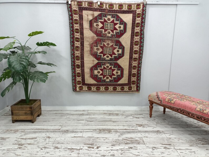 May include: A vintage Turkish rug with a beige background and intricate red, green, and blue geometric patterns. The rug is hanging on a white wall with a wooden floor and a pink and white bench in the foreground.