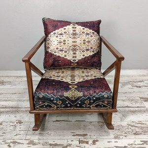 May include: A wooden rocking chair with a patterned cushion and backrest. The cushion and backrest feature a burgundy, cream, and gold design. The chair has a natural wood finish and a classic rocking base.