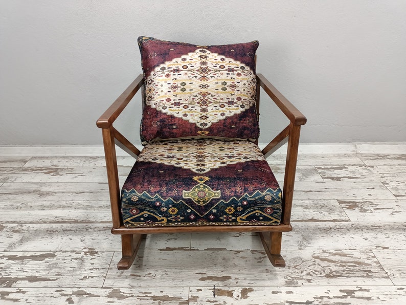 May include: A wooden rocking chair with a patterned cushion and backrest. The cushion and backrest feature a burgundy, cream, and gold design. The chair has a natural wood finish and a classic rocking base.