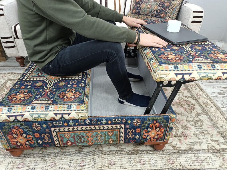 May include: A blue and orange patterned ottoman with a lift-top lid. The ottoman is open and a person is sitting on the lid. A laptop and a white mug are on the top of the ottoman.