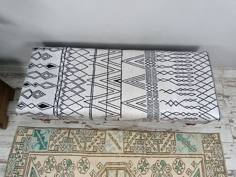 May include: A white and black patterned storage ottoman with a geometric design. The ottoman is sitting on a rug with a similar pattern.