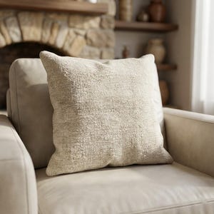 May include: A square, textured beige pillow rests on a light beige armchair. The pillow has a woven appearance and is centered on the chair. The background includes a stone fireplace and wooden shelves, suggesting a cozy living space.