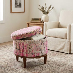 a cozy living room with a pink and white patterned ottoman, a beige armchair, and a wooden side table with a stack of books and a vase of dried flowers.