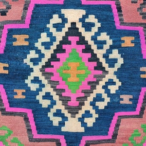 Puede incluir: Un primer plano de una alfombra tejida colorida con un patr&oacute;n geom&eacute;trico en tonos de rosa, azul, verde y marr&oacute;n. La alfombra tiene un dise&ntilde;o tradicional con una forma de diamante central rodeada de detalles intrincados.
