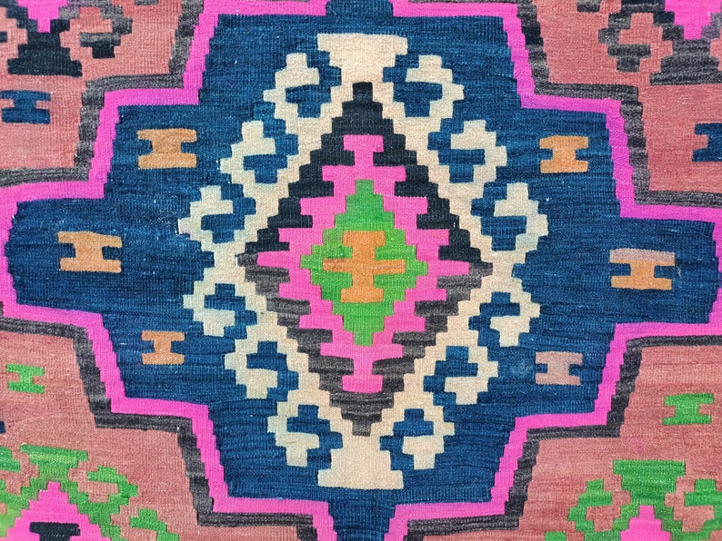 Puede incluir: Un primer plano de una alfombra tejida colorida con un patr&oacute;n geom&eacute;trico en tonos de rosa, azul, verde y marr&oacute;n. La alfombra tiene un dise&ntilde;o tradicional con una forma de diamante central rodeada de detalles intrincados.