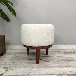 Puede incluir: Otomana redonda blanca con una cubierta de tela texturizada y una base de madera con tres patas.