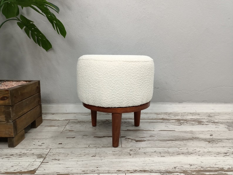 Puede incluir: Otomana redonda blanca con una cubierta de tela texturizada y una base de madera con tres patas.