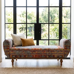 May include: A patterned bench with a wooden frame and curved armrests. The bench features a red, gray, and yellow geometric design. Two pillows, one cream-colored and one mustard-colored, rest on the bench. The bench is placed in front of a large window.