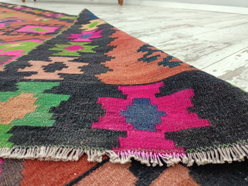 Puede incluir: Primer plano de una alfombra tejida colorida con fondo negro y patrones geom&eacute;tricos. La alfombra tiene un borde de flecos y est&aacute; hecha de lana.