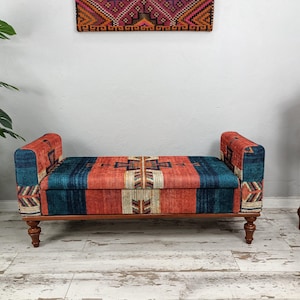 May include: A wooden bench with a colorful, patterned fabric seat and back. The fabric features a design with red, blue, and beige stripes and geometric shapes. The bench has curved armrests and turned wooden legs, adding a touch of elegance.