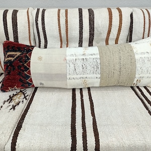 Peut inclure: Un long coussin rectangulaire avec un motif patchwork. Le coussin est fait de différents tissus dans des tons de blanc, beige et marron. Le coussin a une section à motifs rouge et noir à une extrémité.