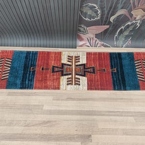 K&ouml;nnte beinhalten: Ein bunter Teppich mit einem geometrischen Muster in Rot-, Blau-, Wei&szlig;- und Braunt&ouml;nen. Der Teppich hat ein tribales Design mit einer zentralen Diamantform und einem Rand aus sich wiederholenden Dreiecken.