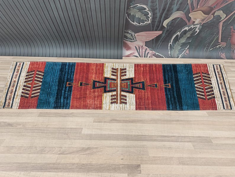 K&ouml;nnte beinhalten: Ein bunter Teppich mit einem geometrischen Muster in Rot-, Blau-, Wei&szlig;- und Braunt&ouml;nen. Der Teppich hat ein tribales Design mit einer zentralen Diamantform und einem Rand aus sich wiederholenden Dreiecken.