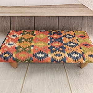 May include: A colourful patterned ottoman with a geometric design in shades of red, orange, yellow, green, and blue. The ottoman has four wooden legs.