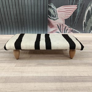 May include: A black and white striped ottoman with wooden legs. The ottoman is made of a soft, woven fabric and has a rectangular shape.