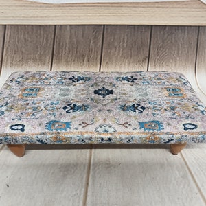 Puede incluir: Un otomano rectangular con una parte superior de tela estampada. La tela es de color beige claro con un diseño floral azul, naranja y marrón. El otomano tiene cuatro patas de madera.